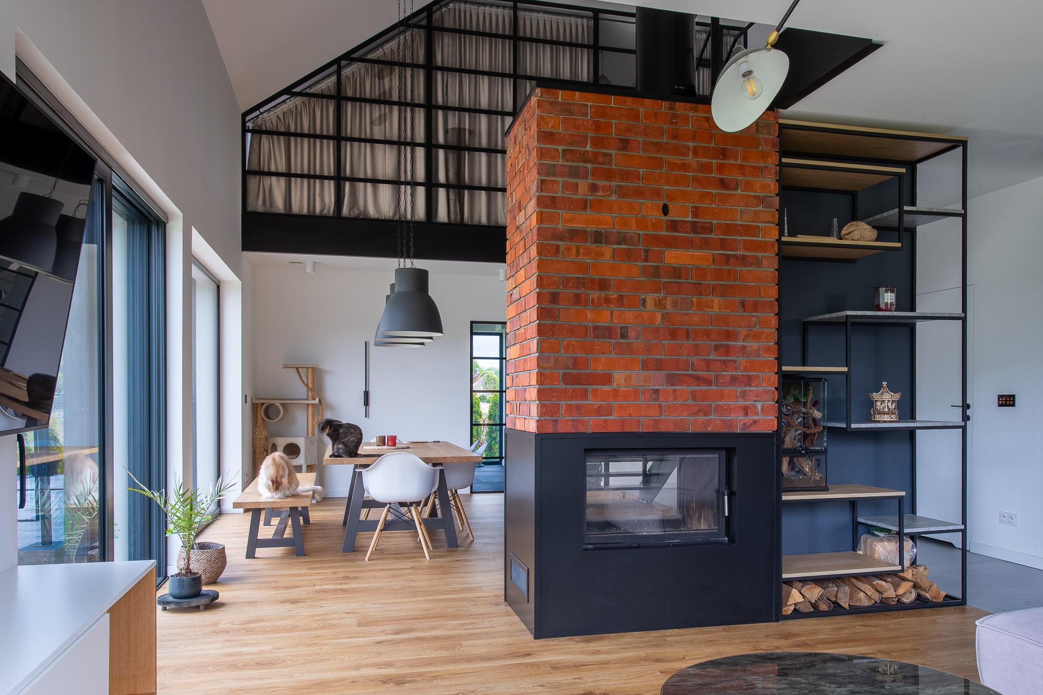 This image shows a modern, open-concept interior space with industrial design elements. The focal point is a striking red brick chimney column that houses a black modern fireplace insert at its base, with a firewood storage compartment below. Adjacent to the brick chimney is a tall black metal shelving unit with glass panels displaying various decorative objects.
The space features light-colored hardwood flooring throughout and high ceilings with exposed black metal framework and curtained panels above. In the dining area, there's a wooden table with modern white molded chairs and a black pendant light hanging above. A person can be seen sitting at the table.
Large glass doors or windows on the left side allow natural light to flood the space, revealing views to the outside. The walls are painted white, creating a bright, airy atmosphere that contrasts with the industrial black metal elements and warm brick tones. A white pendant light fixture hangs from the ceiling near the fireplace area.
The overall aesthetic combines industrial, modern, and minimalist design styles with warm natural materials like wood and brick balanced against sleek metal and glass elements.